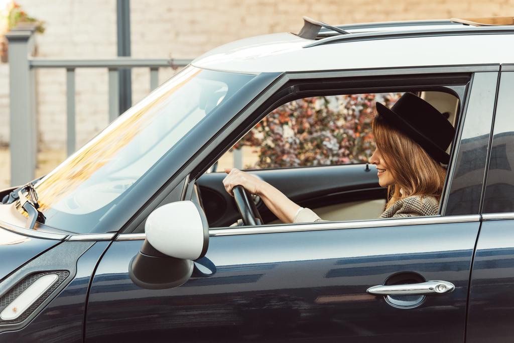 vue latérale de la femme élégante souriante en veste à carreaux et chapeau noir voiture de conduite
  - Photo, image