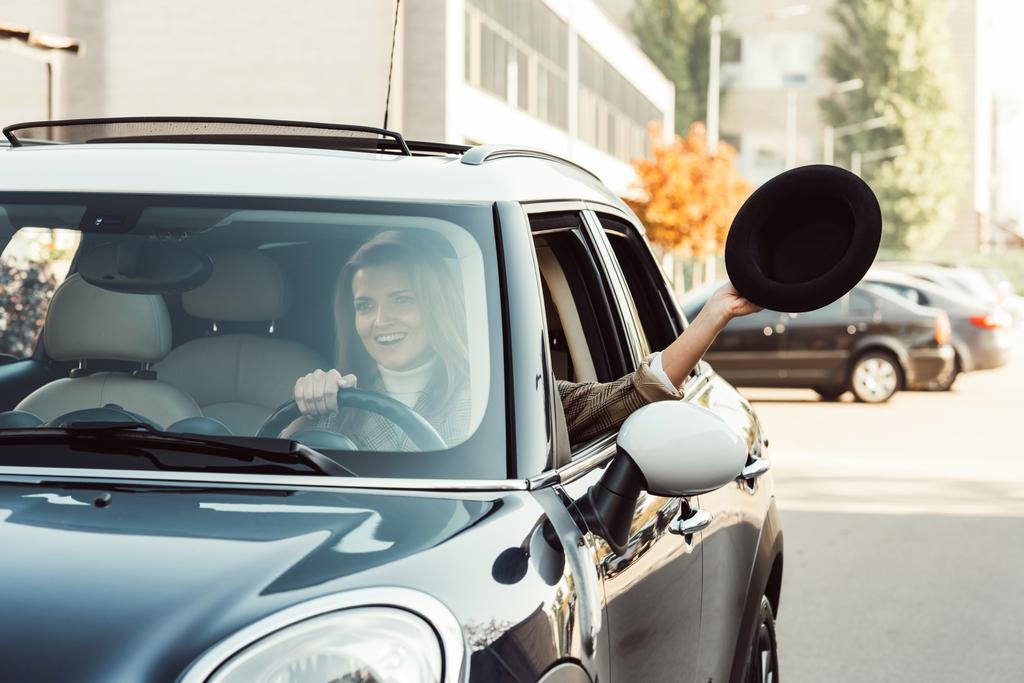 Selektivní fokus veselá žena vyklání ruku s černým kloboukem, zatímco sedí v autě na městské ulici - Fotografie, Obrázek