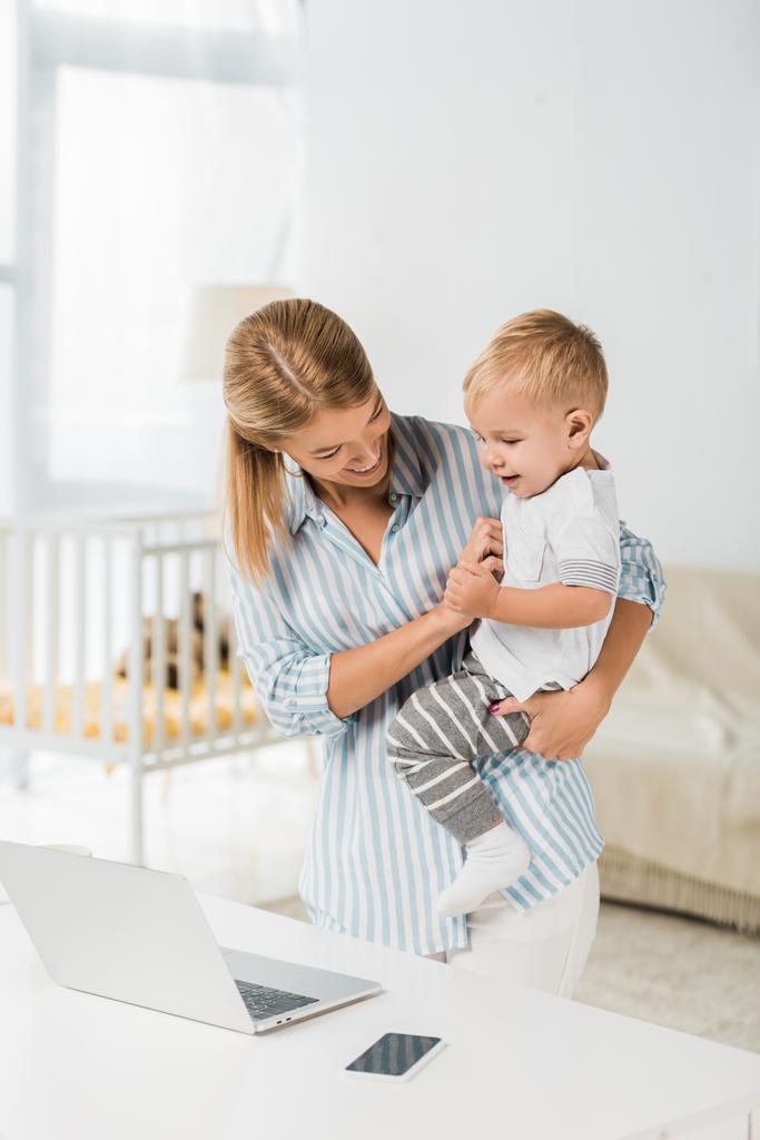 fröhliche Mutter, die mit Laptop und Smartphone am Schreibtisch steht und lächelndes Kleinkind in den Händen hält  - Foto, Bild