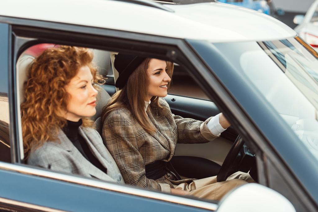 vista lateral da mulher elegante feliz em carro de condução chapéu preto, enquanto seu gengibre amigo feminino sentado perto
  - Foto, Imagem