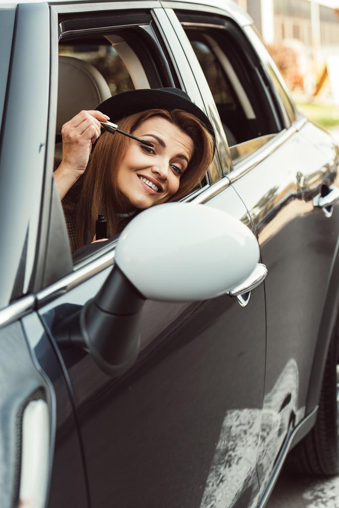 счастливая взрослая женщина в черной шляпе смотрит в зеркало, делая макияж в машине
 - Фото, изображение