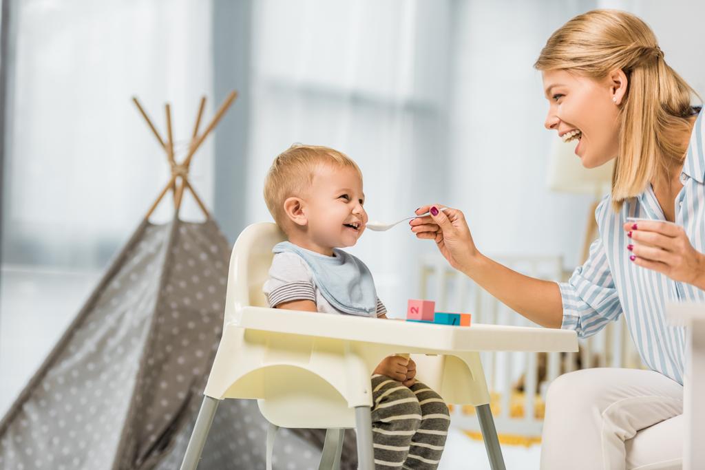 mãe alegre alimentando filho em cadeira alta com comida de bebê no quarto do berçário
 - Foto, Imagem