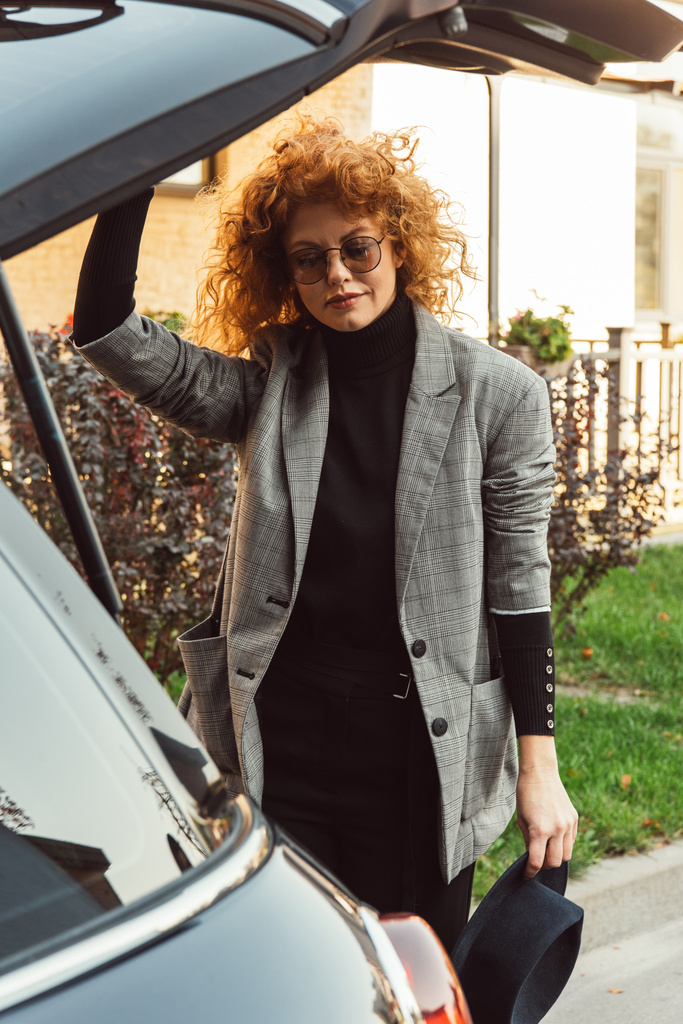 tyylikäs kihara punapää nainen harmaassa takissa ja silmälasit sulkemalla auton runko kaupungin kadulla
 - Valokuva, kuva