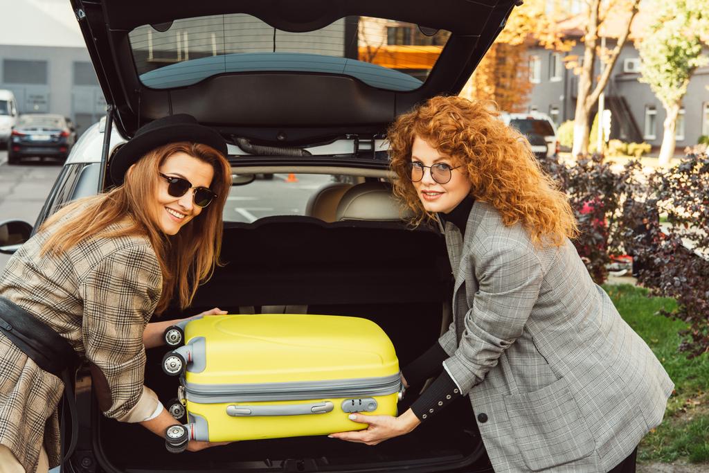 mooie stijlvolle vrouwelijke toeristen in zonnebrillen en jassen zetten wielen tas in de auto kofferbak op stad straat - Foto, afbeelding