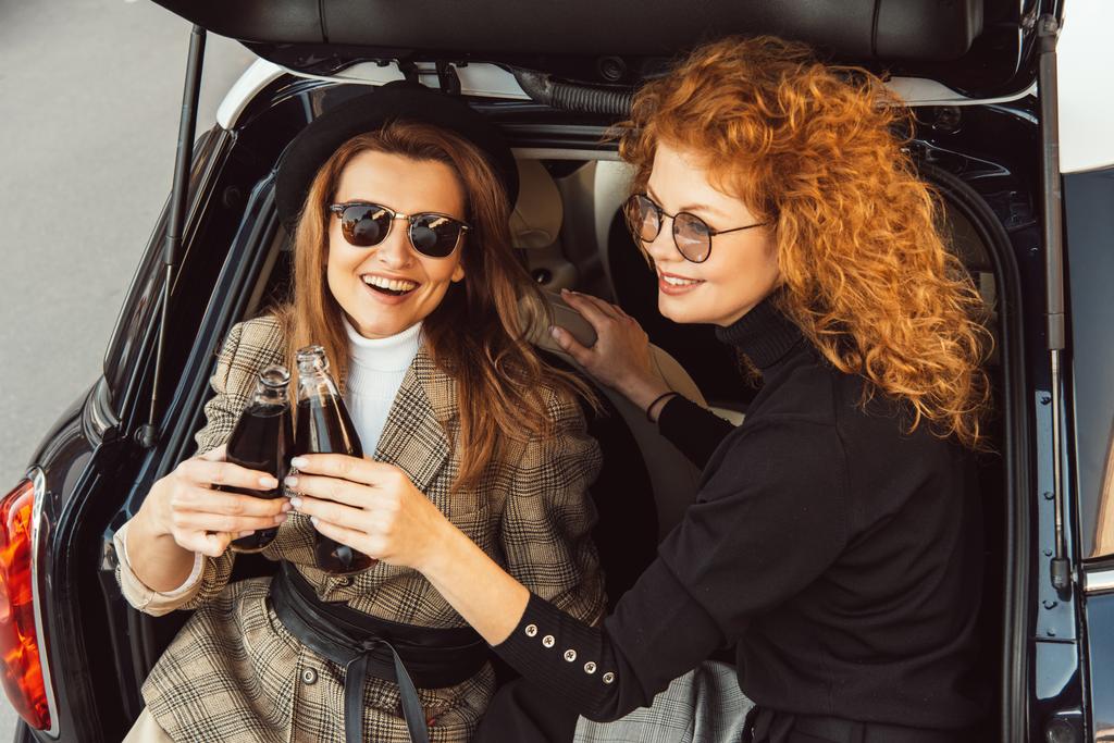hoge hoekmening van glimlachen vriendinnen rammelende door frisdrank flessen in auto kofferbak op stedelijke street - Foto, afbeelding