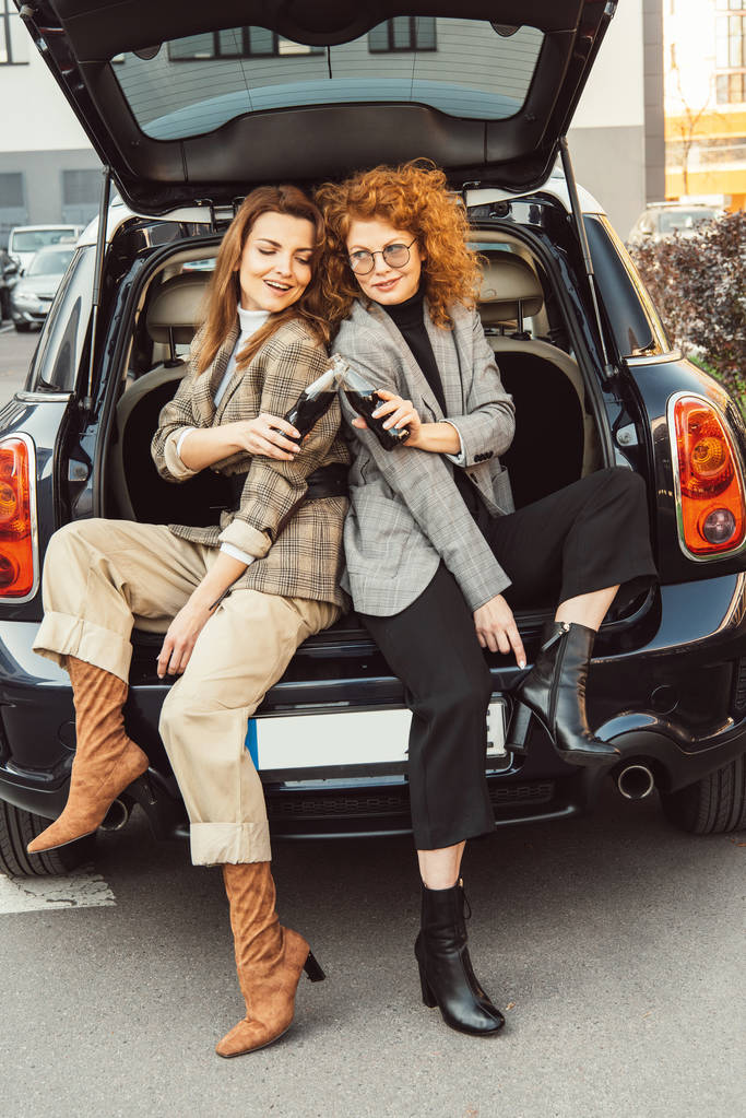 κομψό θηλυκό φίλους σε σακάκια τσούγκριζαν από μπουκάλια σόδας στο πορτ-μπαγκάζ αυτοκινήτου στο δρόμο αστικό - Φωτογραφία, εικόνα