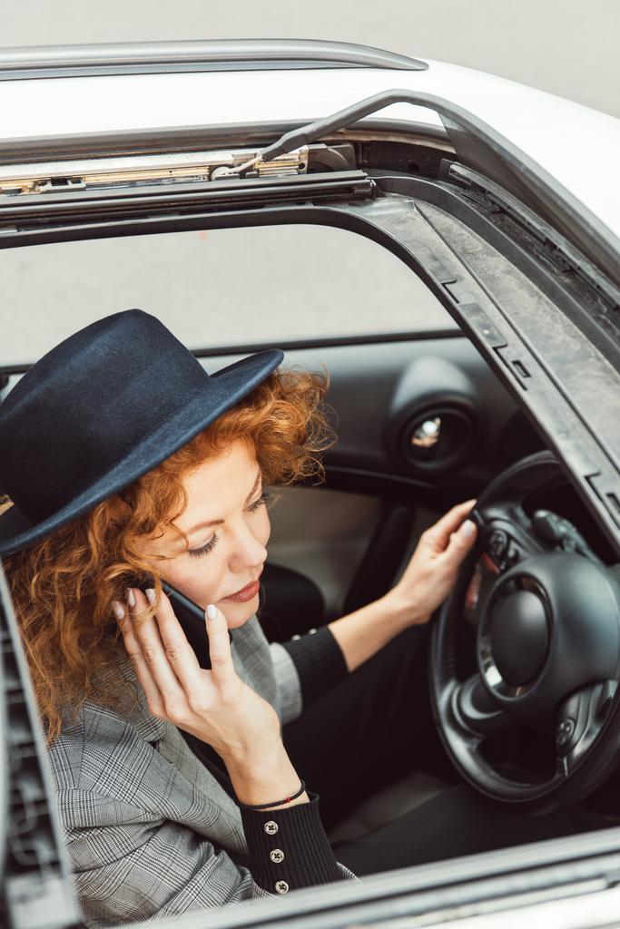 high angle view of redhead woman in black hat talking on smartphone while sitting at steering wheel in car  - Photo, Image