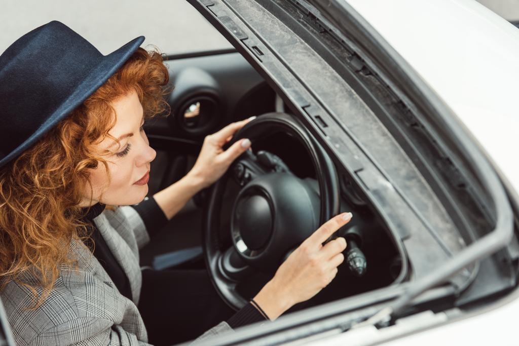 wysoki kąt widzenia modnej kobiety w czarny kapelusz i szara kurtka siedząc przy kierownicy w samochodzie  - Zdjęcie, obraz