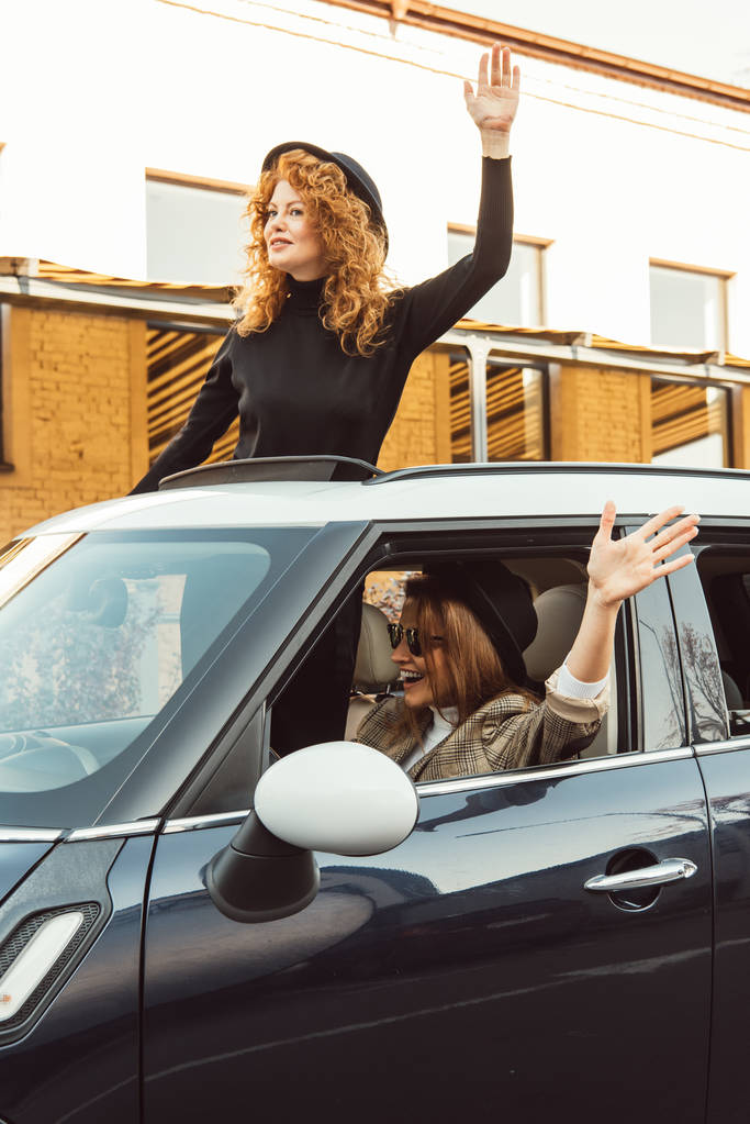 happy female friends in black hats leaning out from car and waving by hands at urban street  - Photo, Image