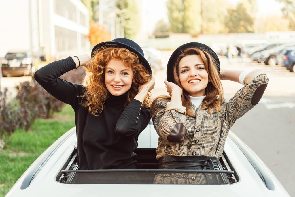 portrait of happy female friends in black hats leaning out from car hatchway at urban street - Photo, Image