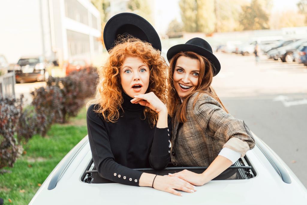 mujer alegre quitándose el sombrero negro de su amiga sorprendida mientras se inclinan hacia fuera de la escotilla del coche en la calle de la ciudad
 - Foto, Imagen