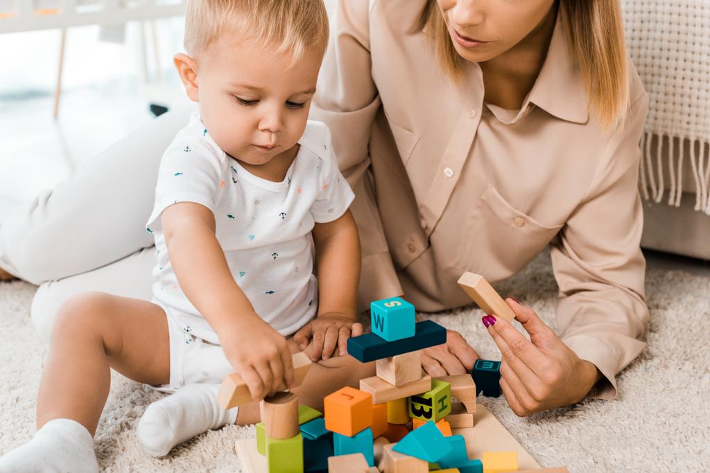 bijgesneden weergave van schattige peuter spelen met kleurrijke kubussen en moeder in de kamer van de kwekerij - Foto, afbeelding