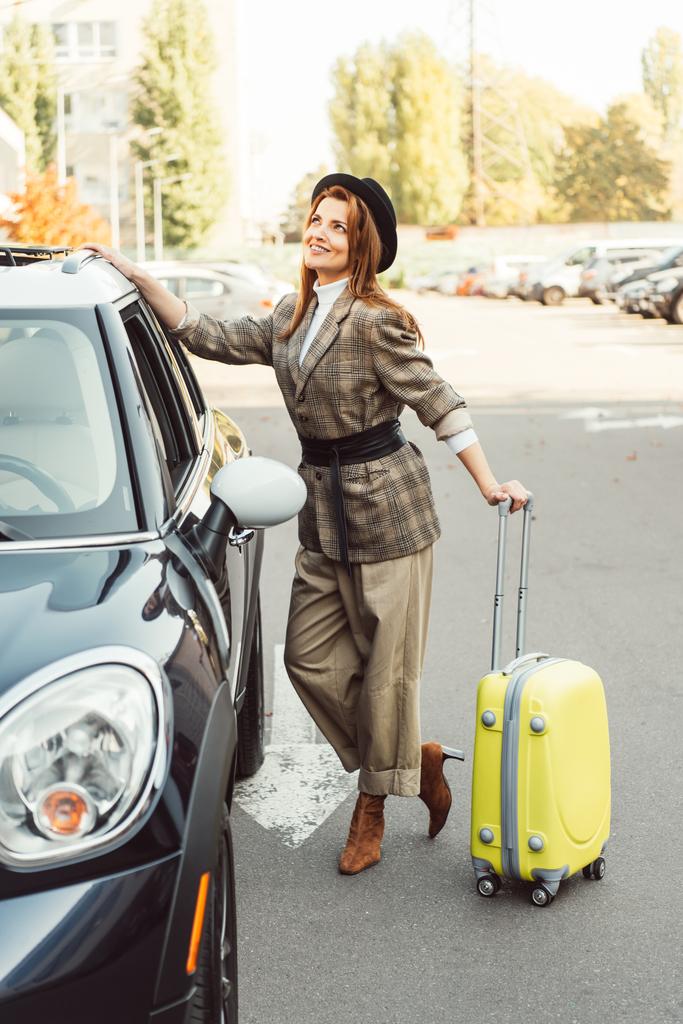 lachende modieuze vrouwelijke toerist in jas staande met wielen tas in de buurt van de auto op straat - Foto, afbeelding