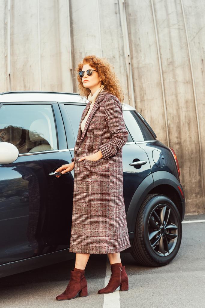 mulher de gengibre encaracolado na moda em casaco e óculos de sol abrindo porta do automóvel na rua da cidade
  - Foto, Imagem