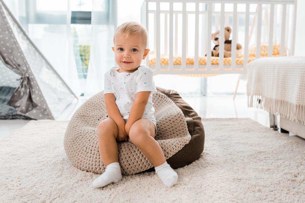 adorable smiling toddler sitting on bean bag chair and looking at camera in nursery room - Photo, Image