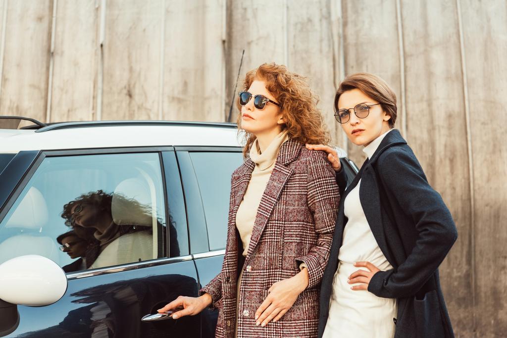 foco seletivo de mulheres adultas elegantes em casacos e óculos de sol posando perto do carro na rua da cidade
 - Foto, Imagem