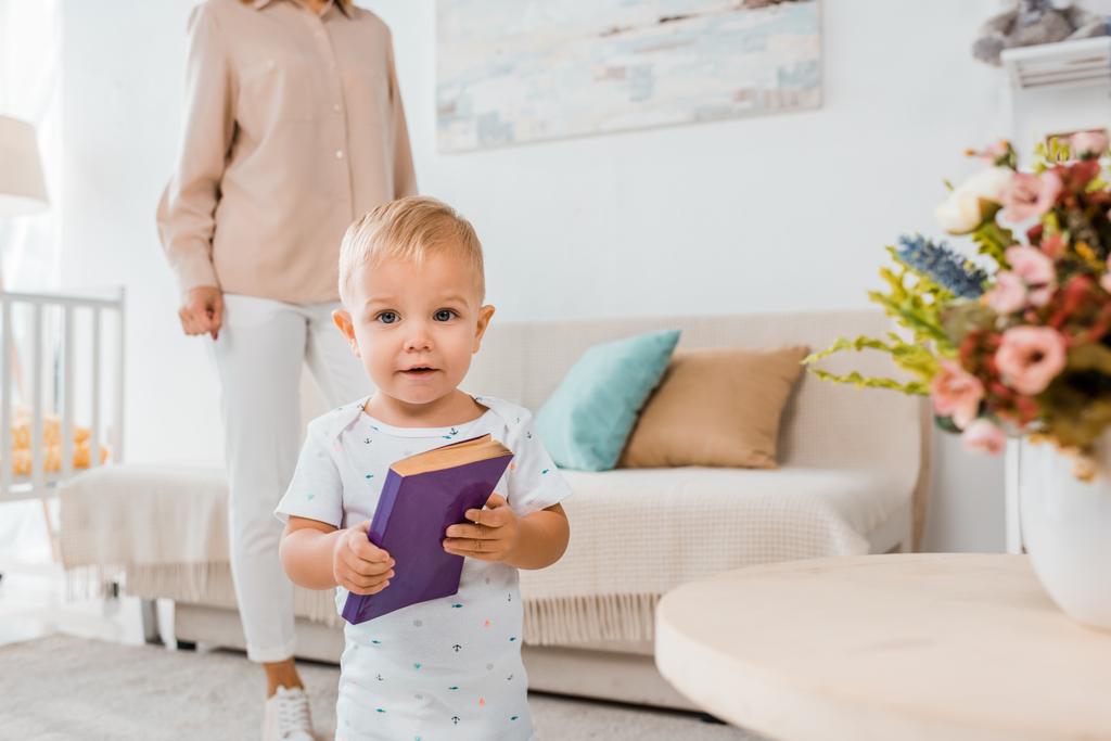 adorable niño sosteniendo libro y mirando a la cámara en el interior con la madre en el fondo
  - Foto, imagen