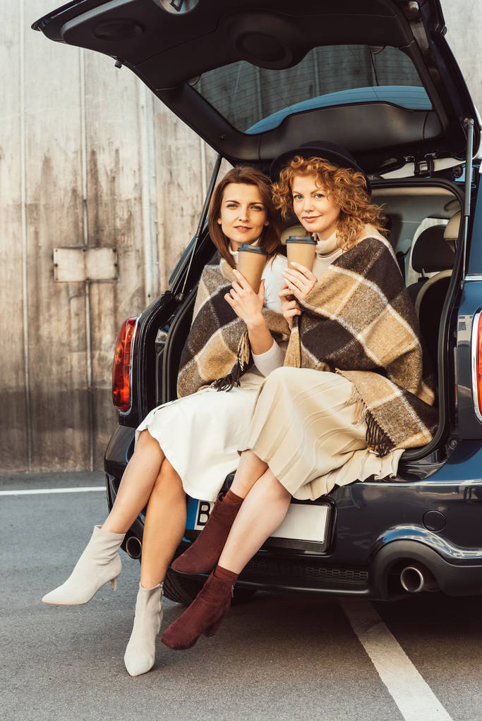 atraentes amigos do sexo feminino envolto em cobertores segurando copos de café descartáveis e sentado no porta-malas do carro na rua da cidade
 - Foto, Imagem