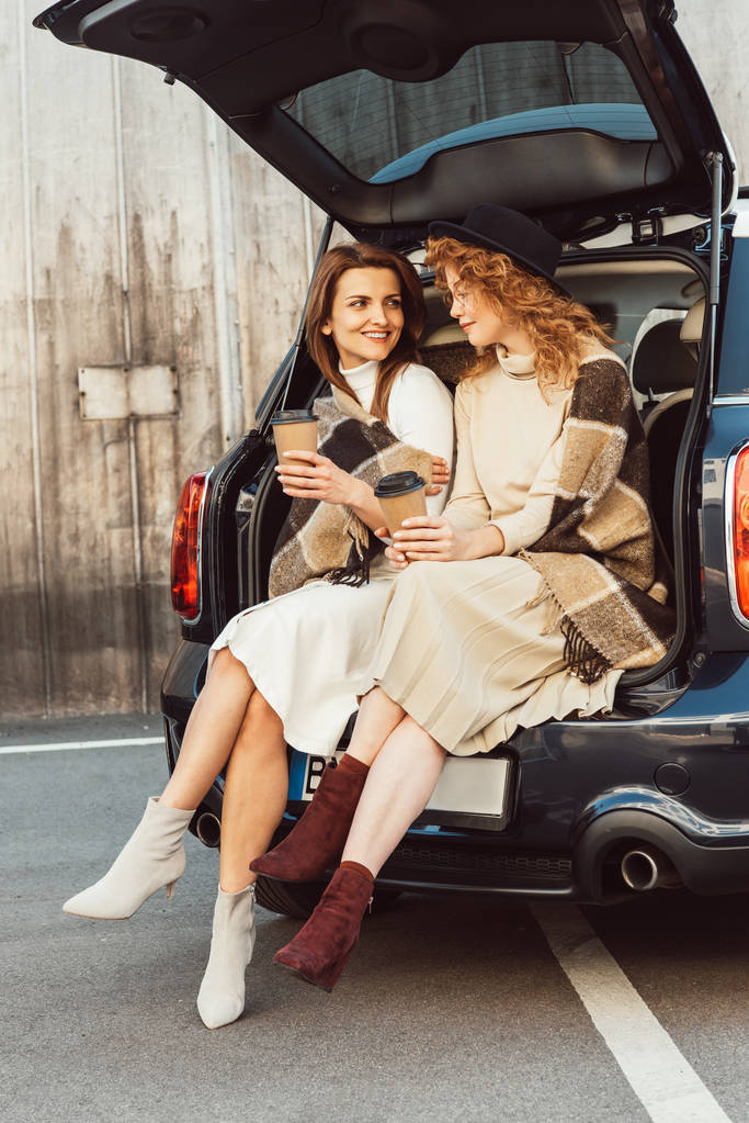 sonrientes amigas sosteniendo tazas de café desechables y hablando mientras están sentadas en el baúl del coche en la calle de la ciudad
 - Foto, imagen
