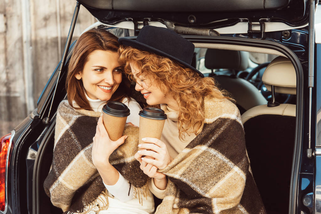 szczęśliwy dorosły koleżanki, owinięte w koce holding Kubki jednorazowe kawy i siedzi w bagażniku samochodu przy ulicy miejskiej - Zdjęcie, obraz