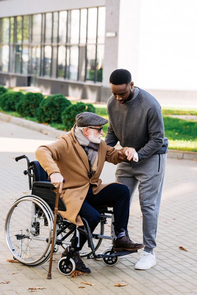 młody Afroamerykanin pielęgniarka pomoc senior niepełnosprawny człowiek wstać z wózka inwalidzkiego na ulicy - Zdjęcie, obraz