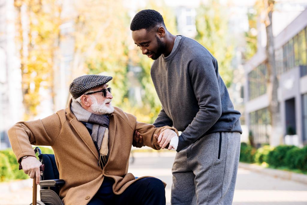 joven africano-americano cuidador ayudando a anciano discapacitado a levantarse de silla de ruedas en la calle
 - Foto, Imagen