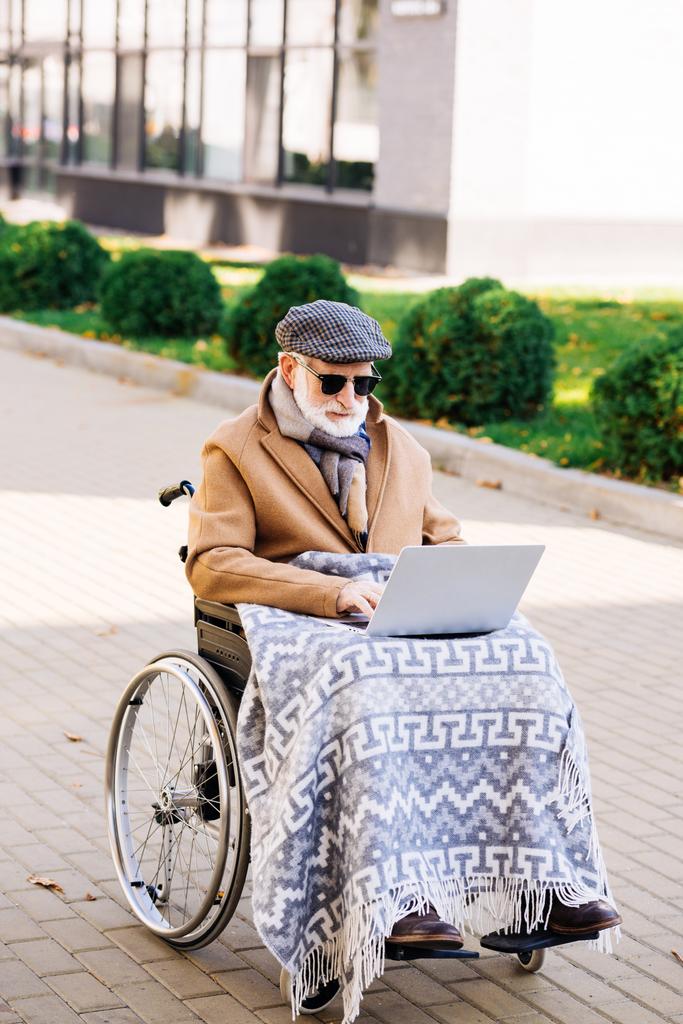 Senior gehandicapte man in rolstoel met plaid werken met de laptop op straat - Foto, afbeelding