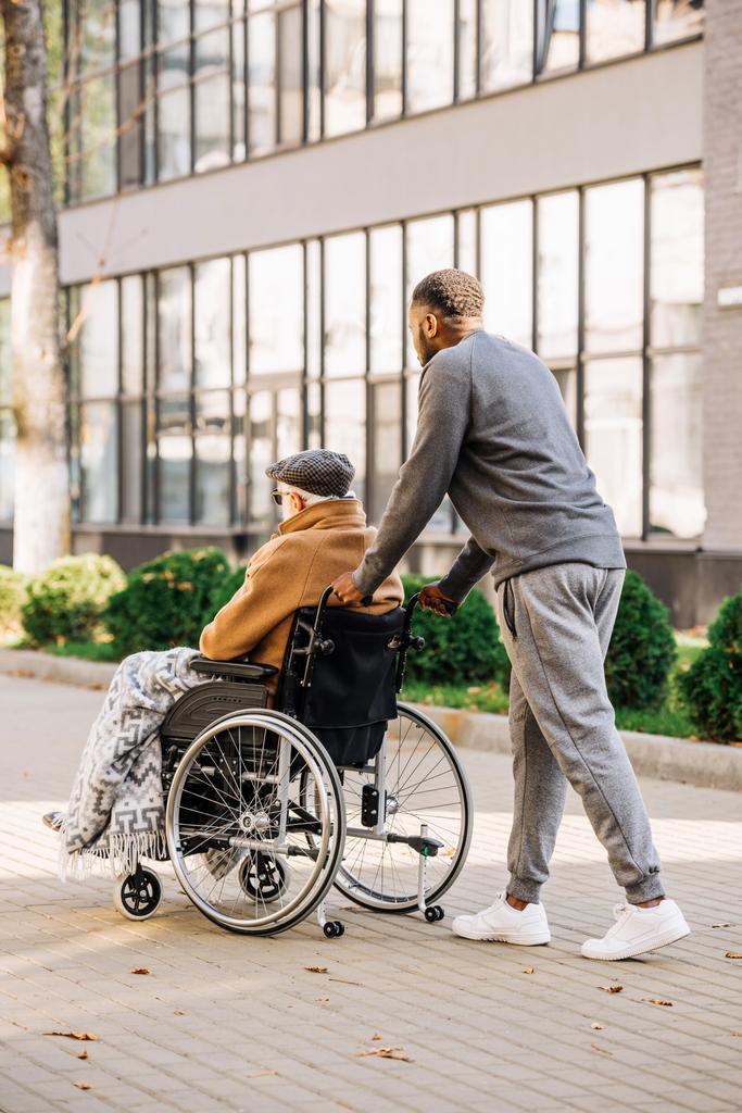 anciano discapacitado en silla de ruedas con cuadros y cuidador cabalgando por calle
 - Foto, Imagen