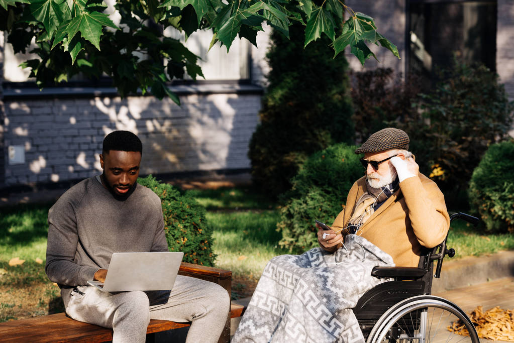 wyłączona starszy człowiek w wózek inwalidzki i młody człowiek african american razem korzystanie z gadżetów na ulicy - Zdjęcie, obraz