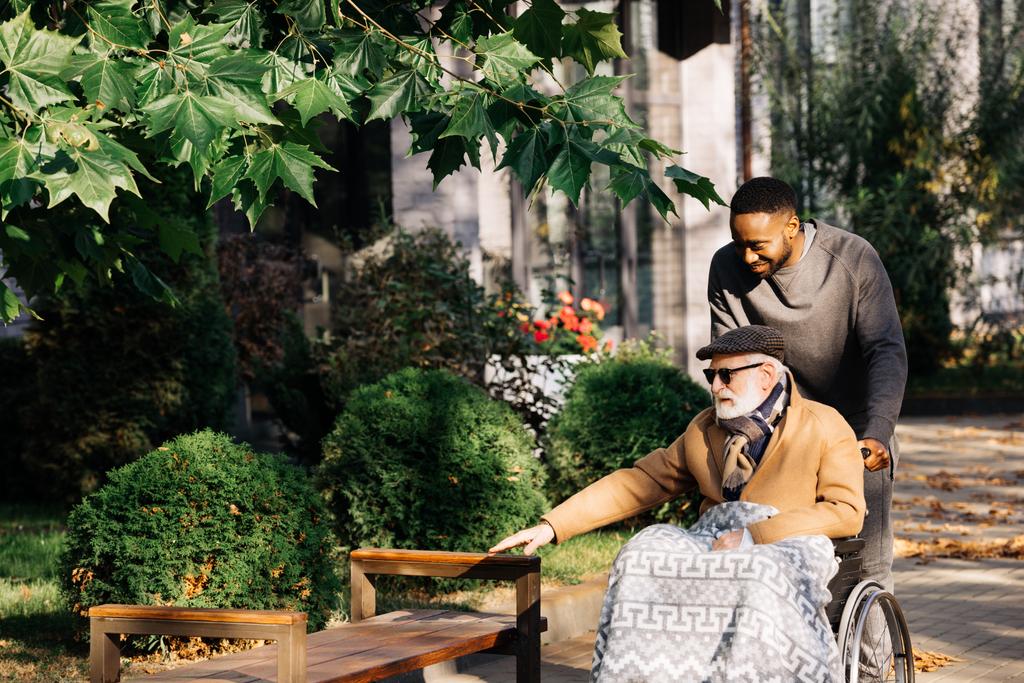 šťastní senioři zakázán člověka do invalidního vozíku a africký Američan cuidador tráví čas spolu na ulici - Fotografie, Obrázek