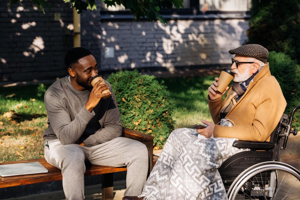 anziano disabile in sedia a rotelle e uomo afroamericano bere caffè da tazze di carta insieme sulla strada
 - Foto, immagini