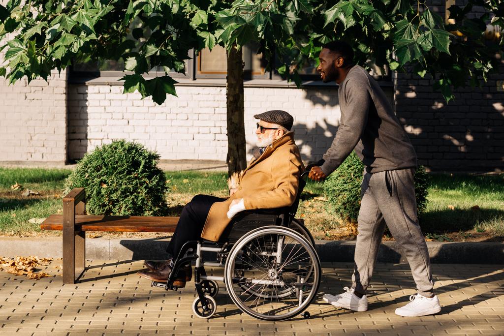 side view of senior disabled man in wheelchair and nurse riding on street - Photo, Image