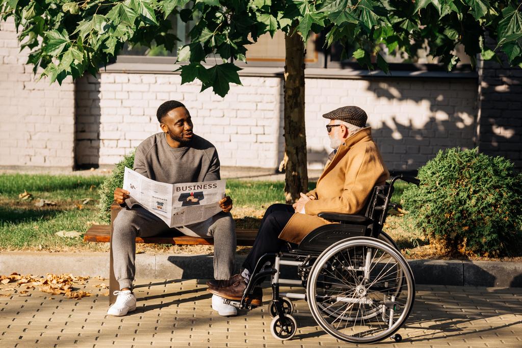 anziano disabile in sedia a rotelle e uomo afroamericano trascorrere del tempo insieme e leggere il giornale per strada
 - Foto, immagini