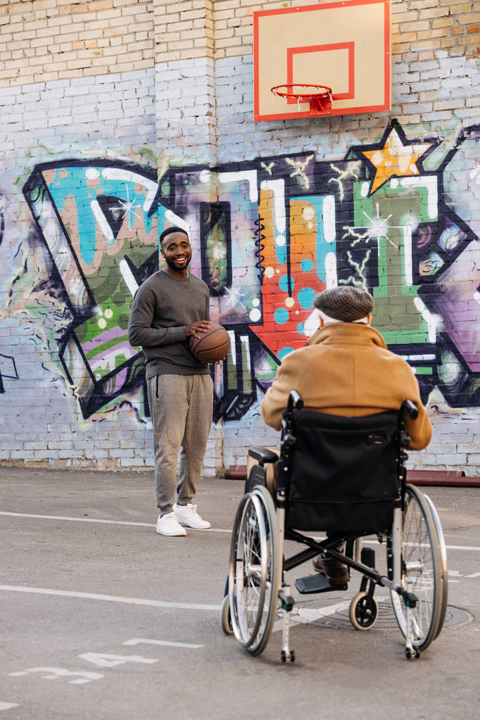 Widok z starszych ludzi niepełnosprawnych na wózku inwalidzkim, patrząc na african american man gry w koszykówkę na ulicy tyłu - Zdjęcie, obraz