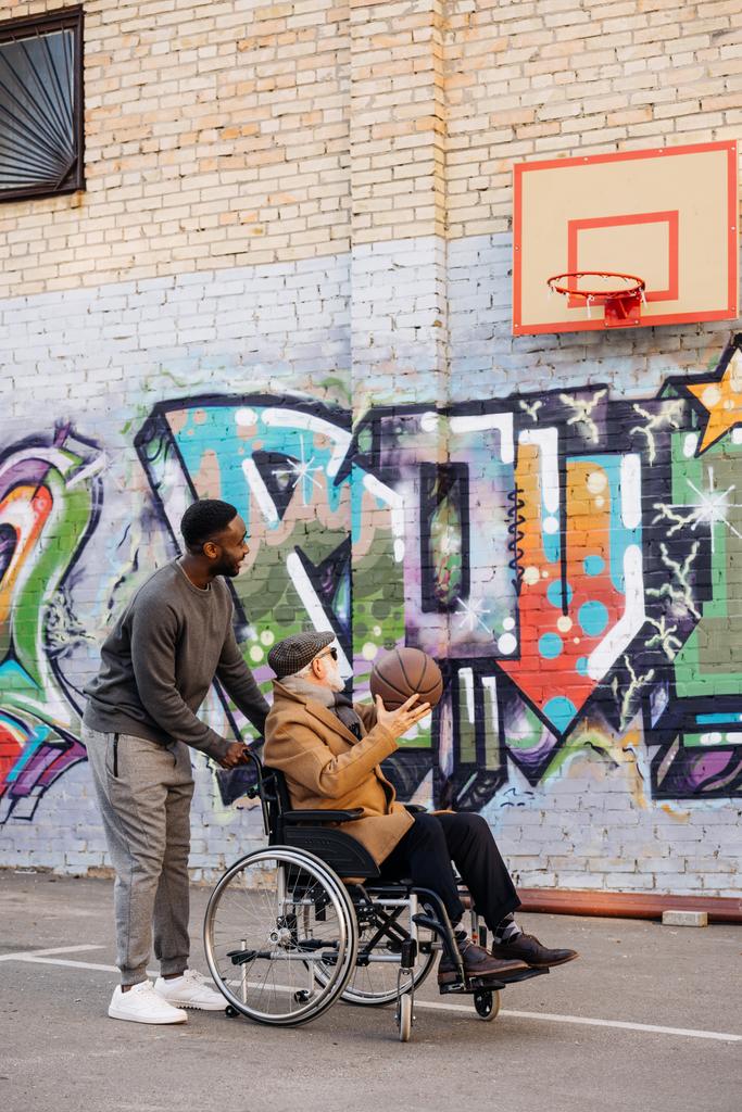 Senior niepełnosprawny człowiek na wózku inwalidzkim i szczęśliwym człowiekiem african american razem gry w koszykówkę na ulicy - Zdjęcie, obraz