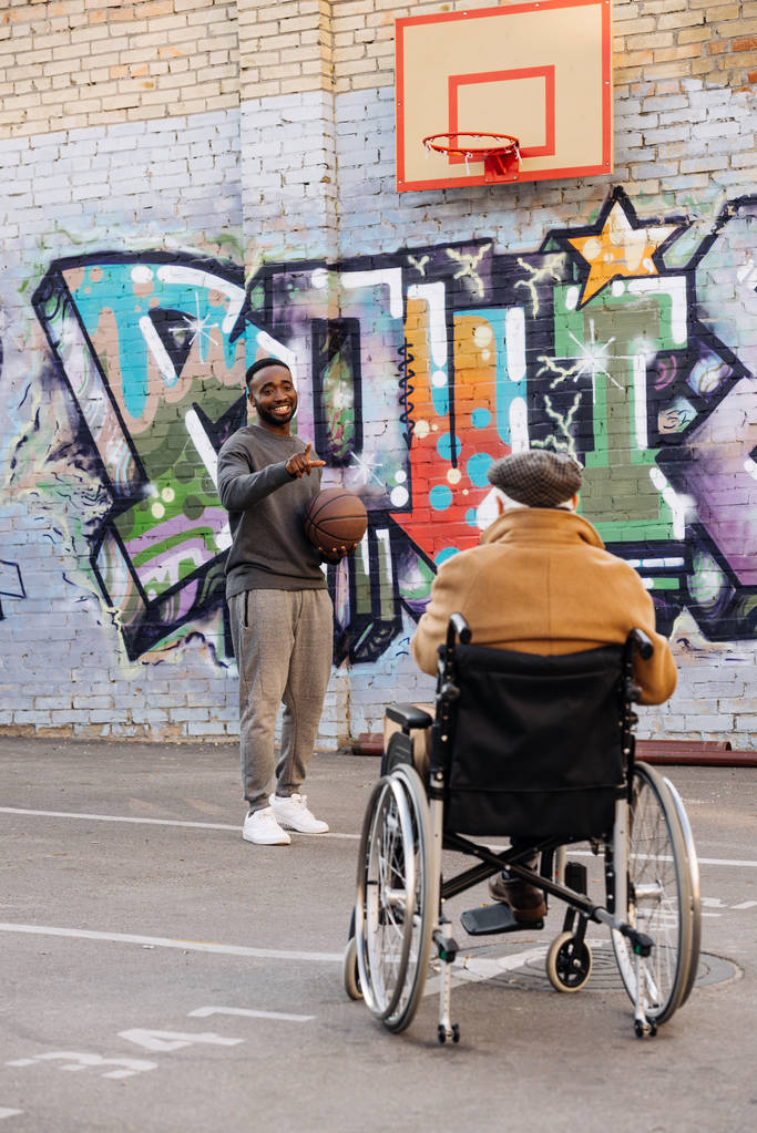 Widok z tyłu z starszych ludzi niepełnosprawnych na wózku inwalidzkim, patrząc na szczęśliwym człowiekiem african american, gry w koszykówkę na ulicy - Zdjęcie, obraz