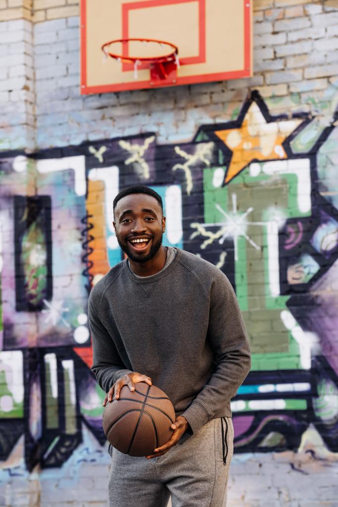 hombre afroamericano feliz con pelota de baloncesto mirando a la cámara en la calle delante de colorido graffiti
 - Foto, Imagen