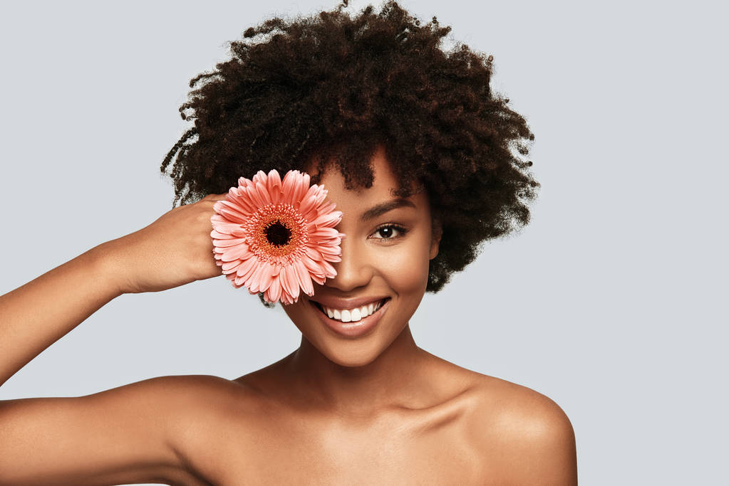 brunette femme africaine regardant la caméra et couvrant l'œil avec des fleurs tout en se tenant sur fond gris
 - Photo, image