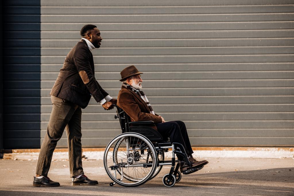 vista laterale dell'uomo anziano disabile in sedia a rotelle e dell'uomo africano americano che cavalca per strada
 - Foto, immagini