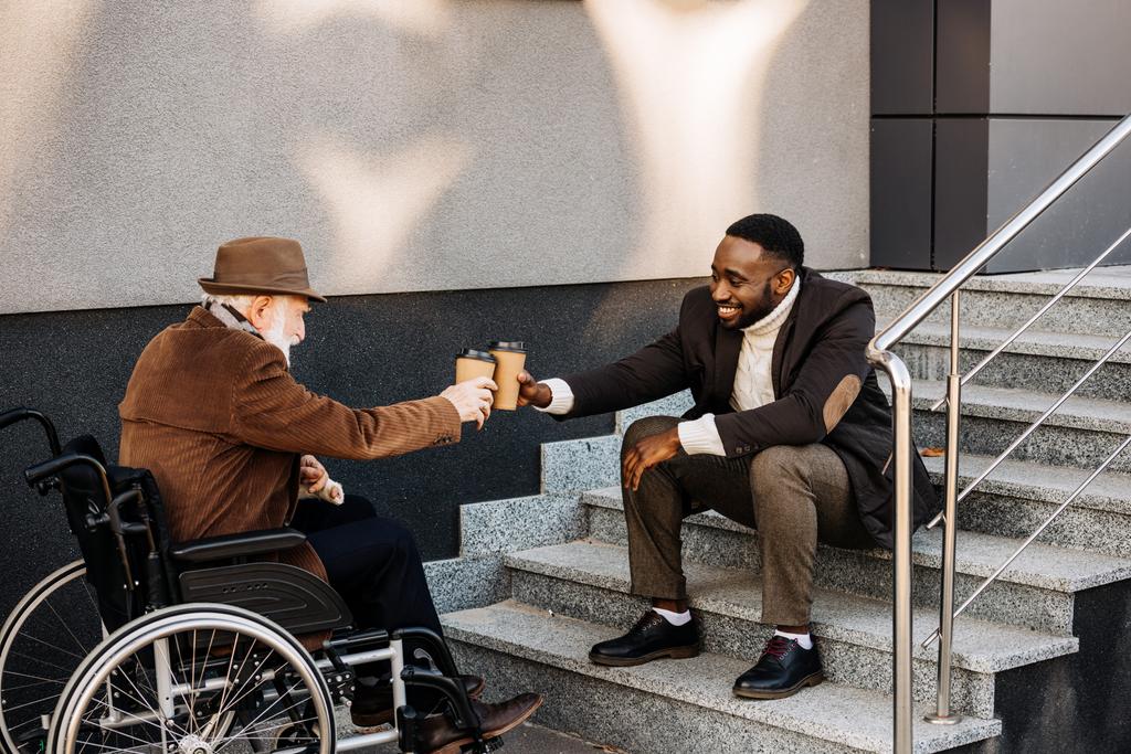 idoso deficiente homem em cadeira de rodas e homem afro-americano bebendo café juntos e copos de papel clinking na rua
 - Foto, Imagem