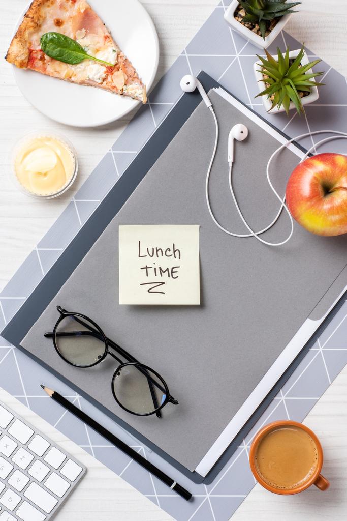 bovenaanzicht van kleverige nota met inscriptie lunchtijd, brillen, koptelefoon, pizza een appel op tafel - Foto, afbeelding