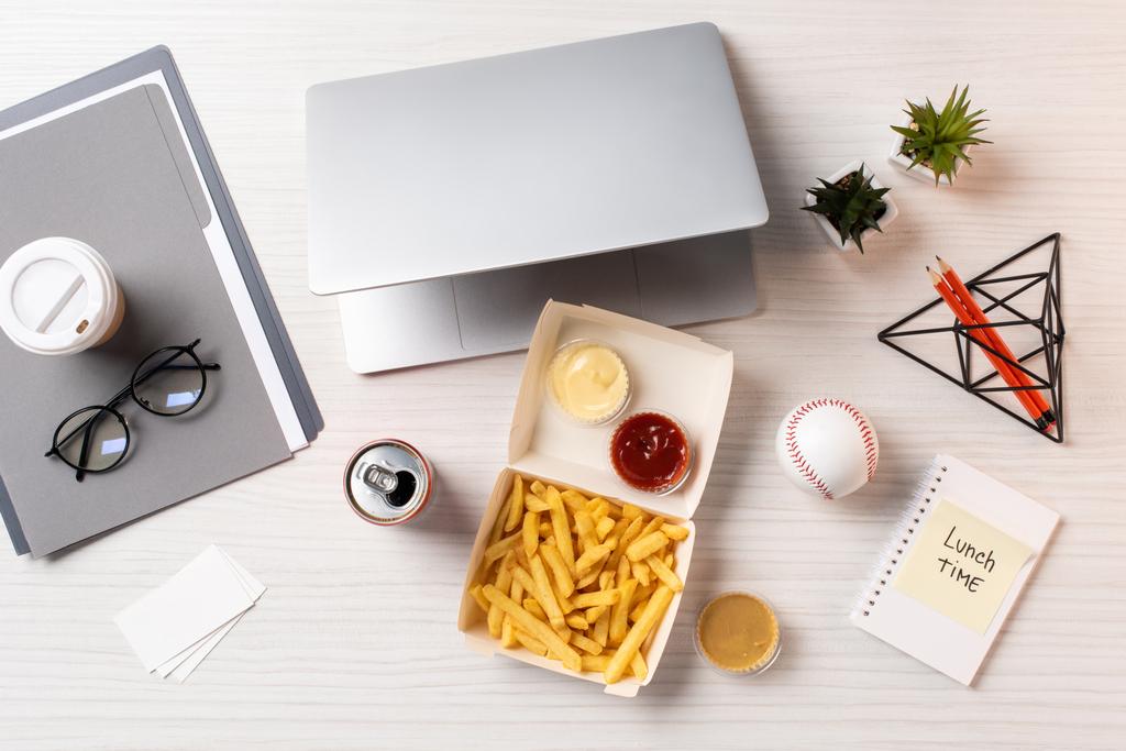 Ansicht von Pommes frites, Getränkedose, Laptop und Bürobedarf am Arbeitsplatz - Foto, Bild