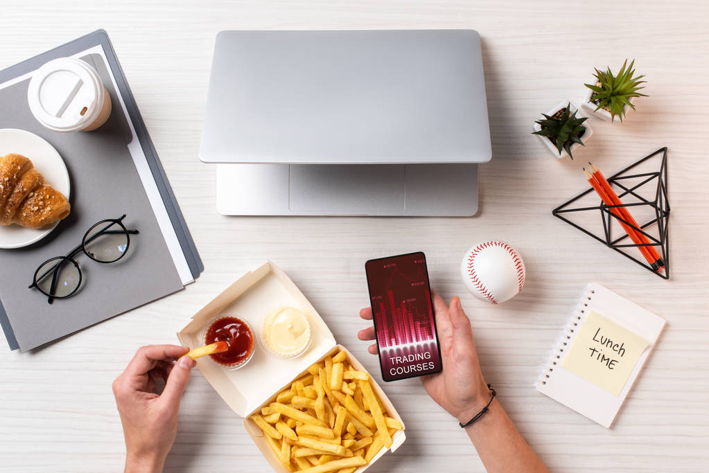 przycięte zdjęcie osoby jedzenia frytek z ketchupem i przy użyciu smartfona z trading kursy w miejscu pracy - Zdjęcie, obraz