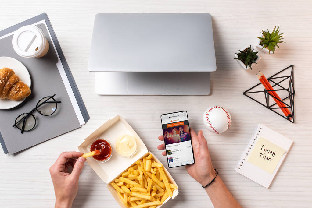 обрізаний знімок людина їсть фрі з кетчупом і за допомогою смартфона з soundcloud app на робочому місці - Фото, зображення