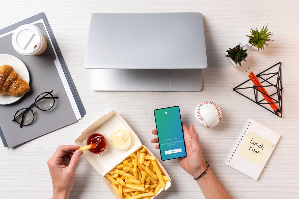 rajattu kuva henkilöstä, joka syö ranskalaisia perunoita ketsupilla ja käyttää älypuhelinta twitter-sovelluksella työpaikalla
 - Valokuva, kuva