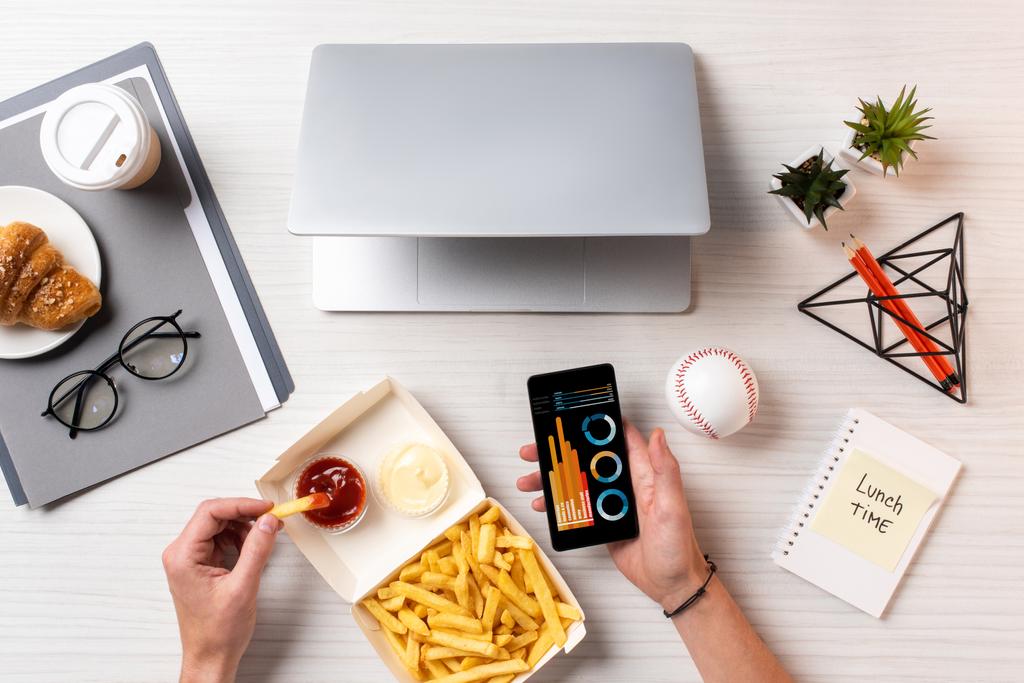 tiro recortado de pessoa comendo batatas fritas com ketchup e usando smartphone com gráficos de negócios no local de trabalho
 - Foto, Imagem