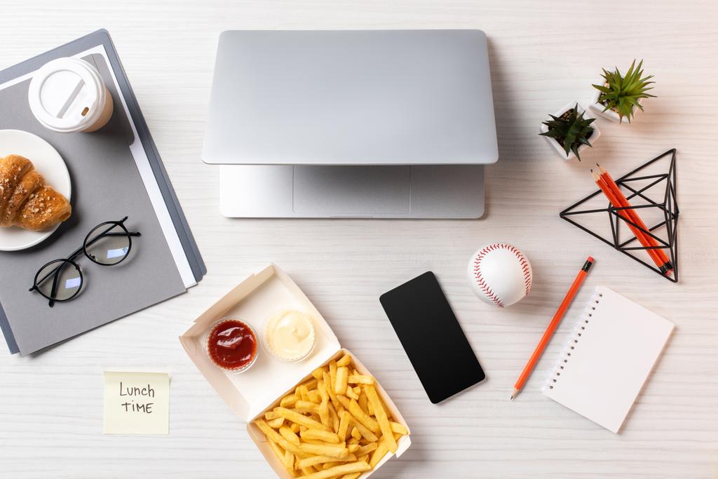 vista dall'alto di patatine fritte, nota adesiva con iscrizione ora di pranzo, laptop e smartphone con schermo bianco sul posto di lavoro
 - Foto, immagini