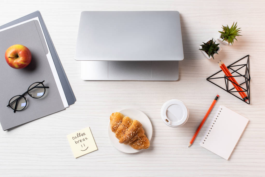 pohled shora na laptop, croissant, jednorázové coffee cup a kancelářské potřeby na stole - Fotografie, Obrázek