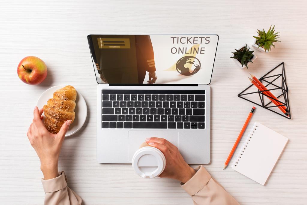 обрезанный снимок предпринимательницы с одноразовой чашкой кофе и круассаном во время использования ноутбука с билетами на сайте
 - Фото, изображение