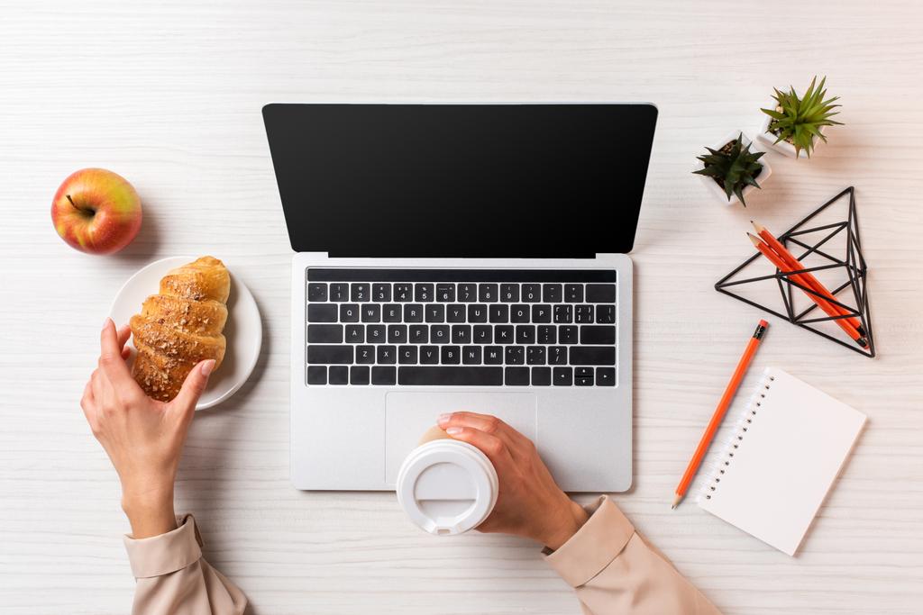 recortado disparo de mujer de negocios sosteniendo taza de café desechable y croissant mientras se utiliza el ordenador portátil con pantalla en blanco
 - Foto, Imagen